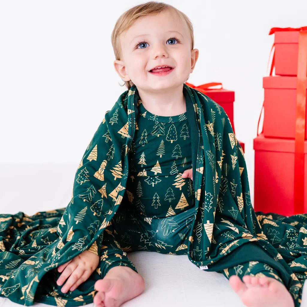 A toddler looking up with a big smile, a dark green blanket in a print of gold and outlined Christmas trees draped around the shoulders, with red gift boxes behind.